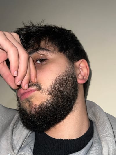 A close-up shot of a person with dark hair and a beard, tilted on its side. The person's right eye is visible and partially obscured by their hand, which is resting on their cheek. A black top is visible at the neck, and a gray hooded sweatshirt is also seen. The background is a plain wall. The person's ear is visible above the hair.