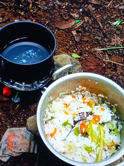 This image captures an outdoor cooking scene in Taungpi, Myanmar (Burma). Two pots are positioned on a rustic, earthy ground, suggesting a campsite or natural setting.
In the upper left, a black pot filled with water sits atop a small, portable camping stove, only partially visible with an orange control knob and metal base. The water appears still, with faint reflections on its surface.
To the right, a larger, silver-colored metal pot contains a cooked meal, likely rice mixed with various vegetables. Visible ingredients include white rice grains, small orange pieces (possibly carrots or squash), green leafy vegetables (like cabbage or spring onions), and some darker, longer pieces that could be mushrooms or other plant matter. A spoon or utensil is partly submerged in the rice mixture.
The ground beneath and around the pots is uneven, dark brown soil, scattered with dry leaves, small rocks, and fragments of what appear to be litter – a green wrapper piece near the top right and a crumpled, discolored piece of paper or plastic with orange markings in the lower left. The overall impression is one of simple, practical outdoor meal preparation. No people are visible, and there is no discernible text. The lighting suggests daytime, possibly overcast or in a shaded area.