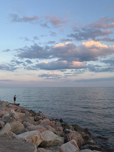 A single individual is fishing on a rocky shore against a backdrop of the ocean and a cloudy sky. The person, wearing a white hat, a grey shirt, and dark shorts, stands on a large rock, holding a fishing rod. The ocean surface displays gentle ripples, reflecting the soft colors of the sky. The sky itself is a gradient of pale blue near the horizon, transitioning to a deeper blue overhead, with scattered clouds tinged with pink and orange from what appears to be sunset. The shore is composed of large, irregularly shaped rocks of various grey and brown hues. The overall scene suggests a peaceful, solitary activity in a natural coastal environment. There is no identifiable location in the city of Unknown, Ukraine.