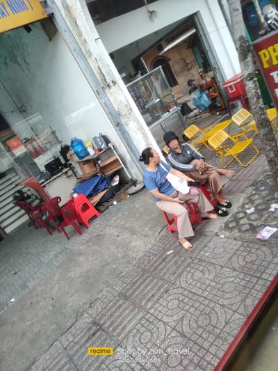 The image depicts a street scene in Thuận An, Vietnam, with two women seated on red plastic stools outside a building. The woman on the left wears a blue t-shirt and light-colored pants, while the woman on the right has on a grey hooded jacket with white stripes on the sleeves and brown pants. She is also wearing a dark hat. In the background, there is a store entrance, a shelf unit with various items, and several yellow plastic chairs placed on the sidewalk. The ground is paved with patterned concrete tiles. A tree trunk is visible on the right side of the frame, and there is some debris on the sidewalk. A red cooler is positioned near the yellow chairs. The overall impression is a snapshot of everyday life on a commercial street.