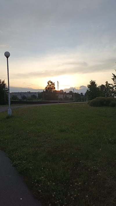 The image depicts a park-like setting with a paved walkway in the foreground, leading towards a grassy field dotted with small white flowers. A tall lamppost stands on the left side of the walkway. In the background, a line of trees separates the park from a residential area with multi-story apartment buildings. The sky is overcast with a warm glow from the setting sun, suggesting it is dusk. The city of Kapyĺ, Belarus is mentioned as the location, implying the scene is within or near this city. There are no people or animals visible in the image.