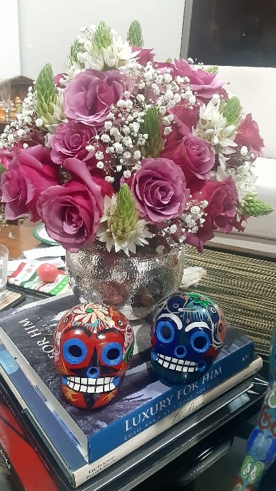 A vibrant bouquet of pink roses, accented by small white flowers and green foliage, dominates the left side. A shimmering silver vase is nestled amongst the blooms. Two painted sugar skulls, one blue and one red, sit atop books with the text "LUXURY FOR HIM" visible. The scene is set against a neutral background, creating a colorful and visually engaging still life.