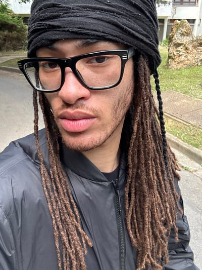 A medium close-up portrait shows a person with brown dreadlocks and a black headwrap, wearing black-rimmed glasses and a dark jacket. The person is partially visible, with the focus on their face. The background includes a sidewalk, grass, trees, and part of a building. The location is Les Ulis, France.