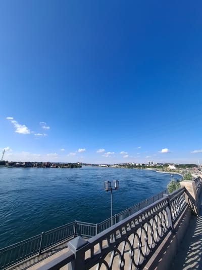 This is a photograph of a wide river in a city. The river is deep blue and appears to be flowing. The sky is clear and bright blue, with a few white clouds scattered across it. On the far bank of the river, there is a line of houses and trees, and in the distance, buildings can be seen, suggesting a city skyline. In the foreground, there is a paved walkway with a decorative metal railing. A lamppost is visible on the walkway. The scene appears to be a waterfront promenade.
Based on the visual elements and information available about Irkutsk, Russia, this scene is likely the Angara River embankment in Irkutsk. The architecture of the buildings on the far bank, as well as the style of the promenade and railing, are consistent with known public spaces in the city. The presence of the wide river and the city skyline are characteristic of Irkutsk's geographical features and urban landscape.