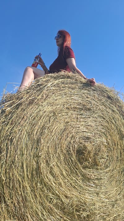 A woman with red hair sits on top of a large, round hay bale, holding a bottle of red liquid. She is wearing a maroon t-shirt and sunglasses. The hay bale is positioned in an outdoor setting with a clear blue sky as the backdrop. The image appears to be taken from a low angle, looking up at the woman and the hay bale. The location is identified as Забалоцкі сельскі Савет, Belarus.