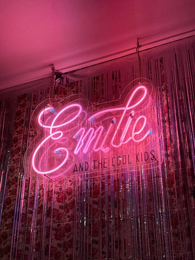 A pink neon sign that says "Lilie" in cursive, with "AND THE COOL KIDS" written to the side in smaller letters. The sign is mounted on a wall with rose-patterned wallpaper behind a shiny, reflective material that hangs in strips. The lighting gives the image a strong pink hue overall. There are cords and hanging mechanisms visible.