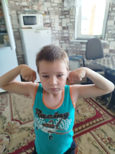 A young boy, with short brown hair and a serious expression, stands in what appears to be a domestic kitchen. He is wearing a turquoise tank top with orange trim that features a racing car graphic and the word "Racing" printed on it. The boy is flexing his biceps, with his arms bent at the elbows and his fists clenched.
The background reveals a kitchen environment with light-colored cabinets and a microwave oven placed on top of a white refrigerator. The walls are covered with patterned wallpaper, and a window with bright daylight is visible. A dark-colored chair is positioned near a table covered with a patterned tablecloth. The floor is carpeted with a floral design. The lighting suggests it is daytime, possibly indoors with natural light contributing to the scene. The overall impression is of a candid or posed photograph of a child showing off his strength.