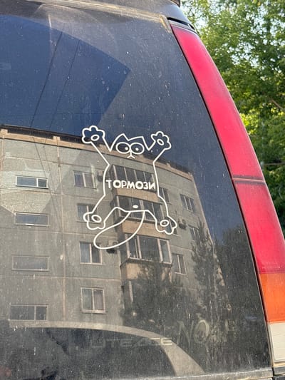 A white cartoon cat sticker is affixed to the rear window of a dirty car. The sticker depicts a cat with wide eyes, outstretched paws, and its body forming a loop. The word "ТОРМОЗИ" (which translates to "Brake" in Russian) is written in the center of the cat's body. In the reflection on the car window, a multi-story apartment building with a beige facade is visible. The building has numerous windows, and a balcony is prominent on the right side. To the right of the window, a red taillight of the car is partially visible. The surrounding environment is out of focus but suggests an outdoor setting with green foliage, likely trees, suggesting the car is parked in a residential area. The presence of the apartment building and the Cyrillic text on the sticker strongly indicates the location is Russia, and potentially Blagoveshchensk given the prompt's constraints.