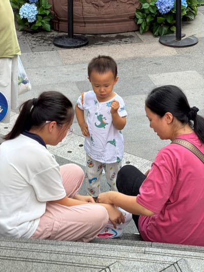 The image shows a scene in Nan'an District, China. Three people are present: an adult woman wearing a pink t-shirt and black pants, an adult woman wearing a white t-shirt and pink pants with glasses, and a young child wearing a white t-shirt with dinosaur prints and patterned pants. The two adult women are sitting on steps, and the child is standing between them. The woman in pink is holding a white object with red and blue designs. The woman in white is looking down, and the child is also looking down. The environment appears to be an outdoor public space, with a paved ground and some potted plants with blue flowers visible in the background. The lighting suggests it is daytime.