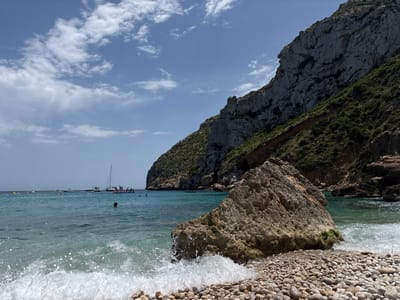 This is a ground-level, eye-level shot of a rocky beach in Xàbia/Jávea, Spain, featuring a cove with calm, turquoise water. In the foreground, a large boulder sits on a shore of smooth, grey and brown pebbles, with a wave breaking against it. The middle ground showcases the clear water, with several boats, including a sailboat, moored in the distance. A few people are visible in the water, swimming and wading, and some are gathered on inflatable structures near the boats. The background is dominated by a steep, grey, rocky cliff face covered in sparse green vegetation. The sky above is a clear blue with scattered white clouds.