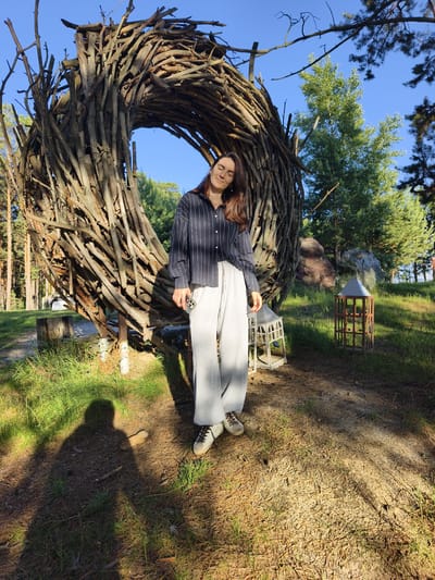 A young woman stands in a sunlit, wooded area in Onochino, Russia, in front of a large, circular sculpture made of intertwined branches. She is wearing a dark, pinstriped button-down shirt and light gray sweatpants, with white and brown sneakers. Her eyes are closed, and she has a relaxed expression. The sculpture is roughly spherical, with an opening in the center, and is constructed from numerous natural branches of varying thickness. Behind the sculpture, the environment consists of green grass, pine trees, and a clear blue sky. Sunlight casts strong shadows across the ground, indicating it is likely late afternoon. In the foreground, her shadow is prominently visible. To the right of the woman, there are two decorative lanterns or birdcages, one white and one wooden. The overall scene suggests an outdoor art installation or park.
