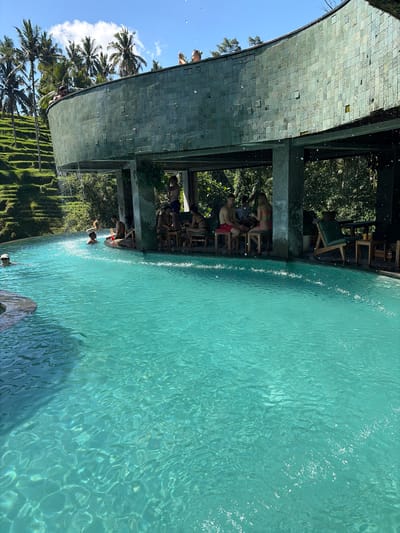 In Tegallalang, Indonesia, a curvilinear structure with a tiled exterior overlooks a turquoise swimming pool. People are visible in and around the pool and under the structure, some swimming, others seated at tables, and some standing. Lush green terraces and palm trees are seen in the background, under a bright blue sky with scattered clouds. The scene suggests a resort or cafe setting by the water.