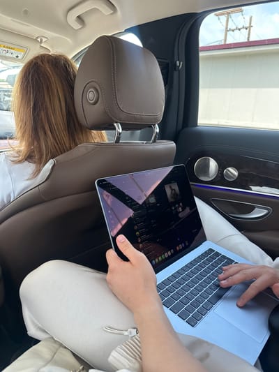 The image captures a daytime scene inside the rear passenger area of a car, viewed as if the camera was rotated 90 degrees clockwise. When viewed upright, it shows two individuals within the vehicle.
In the foreground, a person is seated, with only their hands, lap, and lower torso visible. They are engaged with two electronic devices: an iPad held in their left hand, displaying a digital interface that appears to be a web browser with various icons and tabs; and a light-colored laptop resting on their lap, its QWERTY keyboard clearly visible with standard keys like "control," "option," and "command."
In front of this person, the back of another individual's head and shoulders are seen, indicating they are occupying the front passenger seat. This person has long, light brown hair. The car's interior features luxurious details including brown leather seats with prominent white stitching on the headrest and seat, a dark wood trim on the passenger door, chrome door handles, and two circular speaker grilles, suggesting a premium sound system. A line of blue ambient lighting runs along the door trim.
Through the car's windows, bright daylight is evident, along with a glimpse of the sky and a building or structure. An old wooden utility pole with multiple crossarms and wires stands outside, hinting at a rural or semi-urban environment. A white sun visor on the passenger side displays "AIRBAG" warning text and symbols. The scene depicts activity within a modern vehicle, set in Δημοτική Ενότητα Προμαχώνος, Greece.