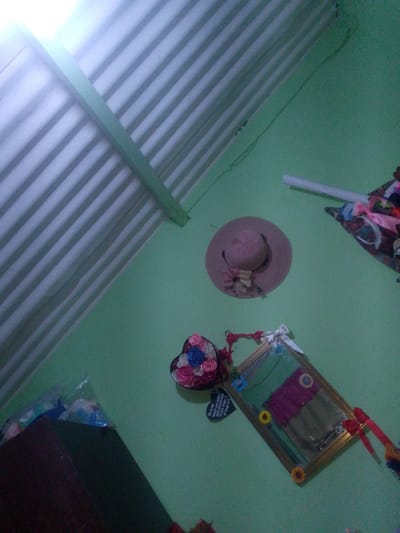 The image shows a room with green walls, likely in El Maco, Venezuela. The ceiling is made of corrugated metal. On the wall, there is a decorative pink hat with flowers, a heart-shaped arrangement of fabric flowers, and a framed mirror with colorful decorations. A white tube light fixture is also visible on the ceiling. To the left, a dark brown cabinet is partially visible, with a plastic bag containing various items on top. The lighting suggests it is daytime, with natural light filtering in from the left, creating shadows on the ceiling. The overall impression is a decorated personal space. There is no discernible activity or event taking place, and no people are visible. The scene appears to be a still life of decorative items in a domestic setting.