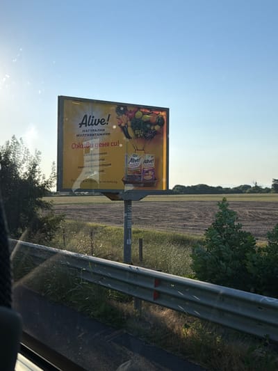 The image captures a billboard advertising "Alive! Natural Multivitamins" in Radinovo, Bulgaria. The billboard features a bright yellow background with the brand name prominently displayed, followed by the text "Natural Multivitamins" and the tagline "Revive your day!". Below the tagline, a list of benefits is presented in Bulgarian: "Strong Immunity," "Healthy Heart," "Memory and Concentration," and "Good Mood." Two product packages of "Alive!" are depicted on the right side of the billboard.
The billboard is situated outdoors, likely along a road, with a rural landscape visible in the background. This landscape includes a harvested field, trees, and a clear blue sky with sunlight casting a bright glare on the left side of the image. A metal guardrail runs along the foreground, obscuring a portion of the lower part of the scene, suggesting the photograph was taken from a moving vehicle. A dark silhouette on the left implies the presence of the photographer or a passenger inside a vehicle. The overall scene is well-lit by natural daylight.