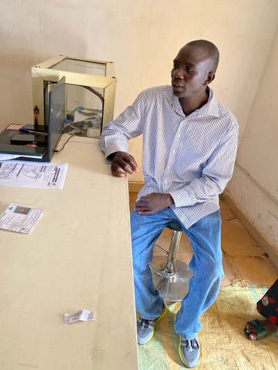 The media file shows a man wearing a blue and white striped long-sleeved shirt and blue jeans, leaning over a counter in what appears to be an office or service center. He has dark skin and is looking down. On the counter, there is a laptop, a file labeled "SIM REPLACEMENT FORM" with "airtel" visible, and other papers. A metallic stool is behind him. In the background, to the right, the feet of another person are visible wearing green sandals, with a floral patterned fabric visible on their clothing. The floor is covered with a yellow, textured mat. The walls are a pale color. The lighting suggests it is daytime. The scene appears to be indoors, likely in Barakin, Nigeria, given the location context. The man's posture and facial expression suggest concentration or engagement with the task at hand.