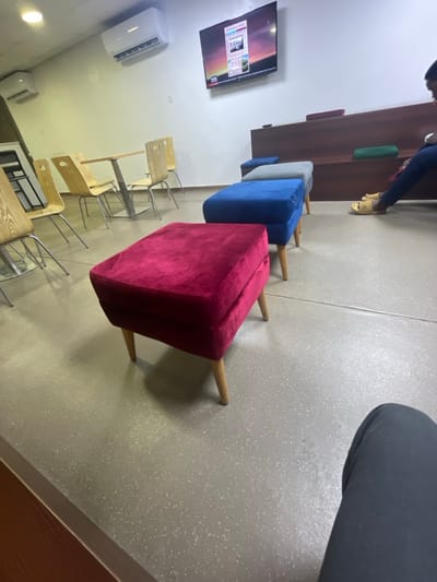 The image displays an indoor waiting or lounge area located in Itirin, Nigeria. The floor is a light gray composite material with white speckles, and the walls are white. Two white wall-mounted air conditioning units are visible on the left wall. A large flat-screen television displaying a news program, with "FINANCIAL TIMES" legible, is mounted on the central wall.

In the foreground, a crimson velvet-upholstered ottoman with light wooden legs is positioned. Behind it, in a linear arrangement, are a blue velvet ottoman and a gray velvet ottoman, followed by a partially visible second blue ottoman, all with similar wooden legs. Along the right wall, a multi-tiered wooden bench structure features green and blue cushions. The lower body of an individual, dressed in dark trousers and light-colored open-toe sandals, is visible seated on the right side of this bench.

In the background on the left, a light-colored rectangular table with a metal base is surrounded by several light wooden chairs with metal frames. Another similar chair is partially visible on the far left. The photograph is taken from a low perspective, with a dark-clad knee, presumably belonging to the photographer, visible in the bottom right corner.