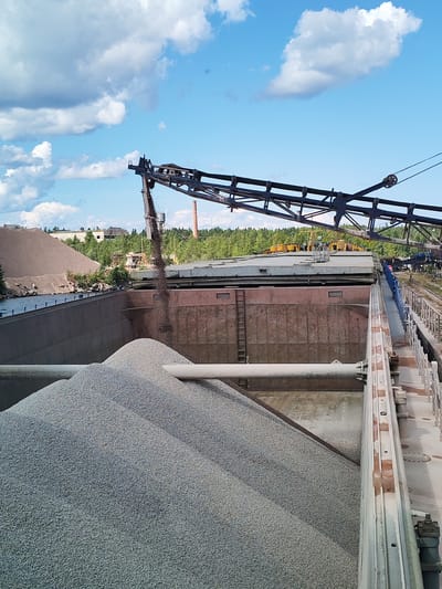 The image depicts an industrial scene in the city of Kvartsitny, Russia, likely at a quarry or processing plant. A large conveyor belt system is actively transferring dark aggregate material from an elevated position into a large, open-topped barge filled with lighter-colored gravel. The barge is positioned alongside a waterway, with a darker brown, rocky embankment visible on the left. In the background, there are large piles of aggregate, industrial structures, trees, and buildings, including a prominent red brick smokestack. The sky is blue with scattered white clouds. The overall impression is one of ongoing material handling and processing in an industrial setting.