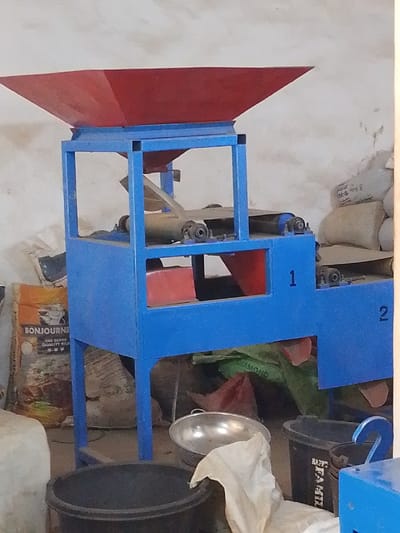 The scene portrays an indoor facility in Olomoda, Nigeria, dominated by a functional-looking industrial processing machine. The machine is constructed with a blue metal frame, supporting a large, red, inverted pyramidal hopper at its uppermost section. Below the hopper, the machine's body integrates various mechanisms, including visible conveyor belts, rollers, and internal red chute components, indicative of a material handling or processing function. The blue frame is sequentially marked with the numbers "1" and "2" on its lower right side.
The machine is positioned against a light-colored, textured wall, likely concrete or plaster, showing minor surface imperfections. The floor, partially visible, appears to be a light, possibly concrete, surface.
Numerous sacks and containers are arranged around the machine. To the immediate left, an orange-brown sack with "BONJOURNE" and "FINE SUPER QUALITY RICE" printed on it is visible. Behind and to the right of the machine, several large, off-white or beige sacks, presumably filled with bulk material, are stacked. A green sack marked "DIAMOND" and a red sack are also present on the floor. In the foreground, multiple black plastic buckets or bins are visible, one of which partially displays "HAPPY FAMIL." A reflective metal bowl, possibly aluminum, rests on the floor, partly obscured by a crumpled white cloth. A white container, possibly plastic, is partially visible on the far left. No individuals are present within the frame, and no actions or interactions are depicted in the static environment.