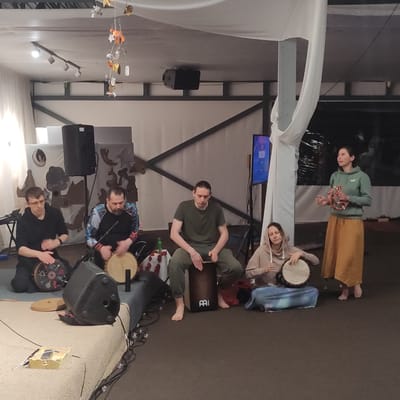 The image captures an indoor gathering where five individuals are engaged in a drumming session. The location is identified as a community space in Zhirnosovo, Russia, characterized by its neutral-colored floor and walls, accented by various decorative elements and lighting fixtures on the ceiling. A white fabric drapes from the ceiling, adding a soft, ethereal quality to the ambiance.

In the foreground, four people are seated or kneeling around drums, actively playing them. The first person on the left, wearing glasses and a black hoodie, is seated and playing a drum adorned with colorful patterns. Next to them, a man in a black, multi-colored patterned shirt is also seated and drumming on a light-colored drum. In the center, a young man with long hair, dressed in a green t-shirt and pants, sits cross-legged and plays a dark brown cajon drum. To his right, a woman in a beige hoodie is seated and playing a hand drum.

Standing on the right side of the frame is a woman wearing a green hoodie and a long, orange skirt. She is holding a tambourine and appears to be leading or participating in the musical activity. A large screen is visible behind her, displaying some sort of content. Speakers are positioned strategically in the scene, suggesting a sound system is in place. The overall scene depicts a relaxed and focused atmosphere, with the participants immersed in their musical activity.