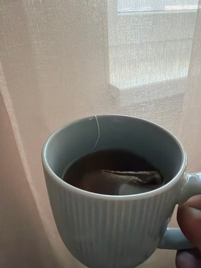 A close-up, eye-level shot shows a hand holding a light blue mug filled with dark tea and a tea bag. The mug has a ribbed texture and a white string hangs from the tea bag. The background is blurred, showing sheer, light pink curtains and a window with a grid pattern. The scene is likely indoors, with diffused light coming from the window. No specific location in Novorossiysk, Russia can be identified from the image.