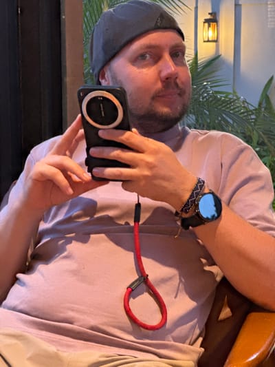 In Nam Hoa Lư Ward, Vietnam, a man wearing a gray beanie and a pink t-shirt is holding a smartphone with a red lanyard. He is also wearing a wristwatch with a black band and a patterned bracelet. The man is sitting in a chair and is looking towards the camera. In the background, there is a wall with a lantern and some palm trees. The lighting is dim, suggesting it is evening or indoors. The man appears to be relaxing or waiting.