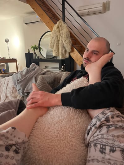 A bald man in a black hoodie sits on a white furry pillow. He holds a foot, likely of another person, near his face, appearing to be playfully annoyed or disgusted. The background shows a modern interior with wooden beams, a staircase with a fur coat draped over it, a round mirror, and an air conditioning unit mounted high on the wall. Another person is lying to the right wearing pajamas.