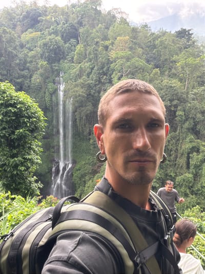 A man with fair skin and short, light-colored hair poses for a selfie with a waterfall and lush green foliage in the background. He wears decorative earrings and a backpack. Two other individuals are faintly visible behind him, one wearing a gray shirt and sunglasses. The lighting is slightly overcast, creating a soft, diffused effect.