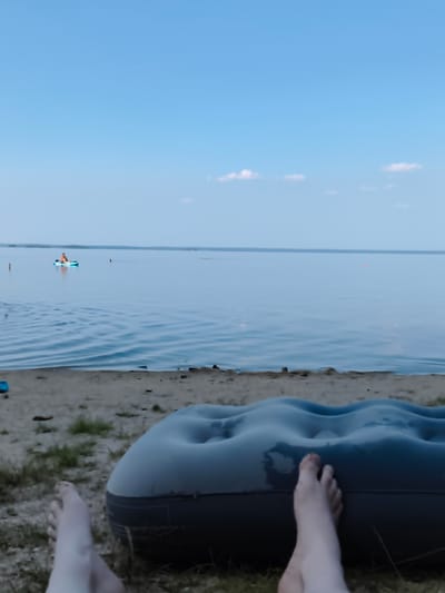 This image depicts a tranquil scene at a body of water, likely a lake or reservoir, with the sky overhead. In the foreground, a person's legs and feet are visible, resting on the ground next to an inflated air mattress. The air mattress is dark gray and appears slightly damp. The ground is a mix of sand and sparse grass.

Beyond the immediate foreground, the water stretches out, calm with gentle ripples. In the middle distance, a person is seated on a small, light blue watercraft, possibly a kayak or paddleboard, on the water. Further out, a faint line of orange buoys is visible, suggesting a designated swimming or boating area. The horizon is marked by a distant shoreline with a faint, hazy landmass. The sky is a clear, pale blue with a few wispy white clouds scattered across it.

The overall impression is one of relaxation and outdoor leisure, with the individual enjoying the peaceful atmosphere by the water. While the prompt requests a location in Krasny Kamen, Russia, there are no specific visual cues in the image to confirm this geographical context. The scene is generic and could be found at many similar natural locations.