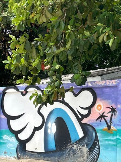 The image shows a colorful mural painted on a wall in Ban Tai Subdistrict, Thailand. The mural features a large cartoon character with white, wing-like shapes on its head and a prominent blue eye with white highlights. The background of the mural depicts a tropical scene with a purple and pink sky, a small island with two palm trees, and blue water with white waves. Trees with green leaves hang over the top of the mural, partially obscuring the wall and roofline above. The wall itself appears to be weathered, with some areas of paint peeling. The text "POPEYE" is visible in pink lettering on the upper part of the wall.