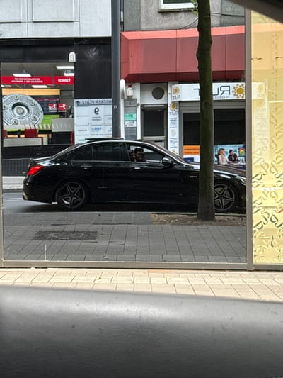 The image captures a street scene in Essen, Germany, likely seen as a reflection in a window or mirror, given the mirrored text on various signs. The primary subject is a black four-door sedan, possibly a Mercedes-Benz, parked parallel to a sidewalk on the right side of the frame. The car features distinctive star-spoke alloy wheels. A person's head is visible through the front driver's side window.
Behind the car, several buildings line the street, featuring various storefronts and architectural details. A thick tree trunk or large branch extends horizontally across the middle of the frame, partially obscuring the background. The sidewalk next to the car is paved with light grey rectangular stones, and a darker grey street or parking area is visible beyond it. A circular, textured metal cover is embedded in the street surface.
Visible text on storefront signs, corrected for mirroring, includes:
* "TÜREN ENERGIE" (Doors Energy) with a sun logo.
* "STAB BECKER"
* A red sign with "fiboco" and text below it stating "ERNEUERBARE ENERGIE UND IT GRENZENLOS" (Renewable Energy and IT Unlimited) and "QUALIFIZIERTES IMPRESSUM" (Qualified Imprint).
The scene appears to be taking place during the daytime, with ample natural light suggesting clear or partly cloudy weather. No specific activity beyond parking is depicted, presenting a static urban snapshot. No particular emotions are discernible.