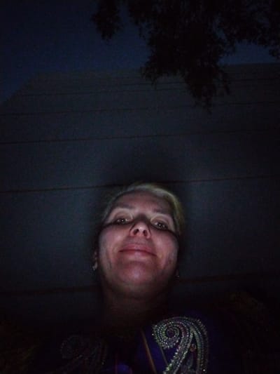 A low-angle, dark, outdoor selfie shows a woman's face and upper chest. The woman has fair skin, blonde hair, and is wearing a dark, embellished top. She appears to be looking up at the camera. The background is dark, with horizontal lines visible, suggesting a wall or siding. A tree branch is visible in the upper portion of the image. The image was taken at night in Al Mas'oudi, United Arab Emirates.