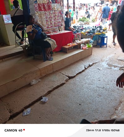 A bustling marketplace scene is captured with vendors displaying wares, including jewelry, shoes, and produce. A woman in a blue shirt sits and appears to be eating. In the foreground, the concrete ground is visibly cracked. People are moving through the market. The scene is lit with natural light. A second woman sits behind a market display of jewelry. A man stands to the left near a display of watches.