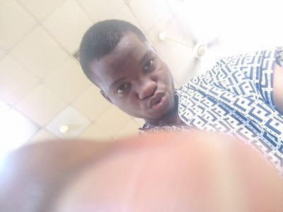 The image is a brightly lit, low-angle selfie taken indoors in Ipetumodu, Nigeria. The primary subject is a young person, likely male, with dark skin and short dark hair, looking directly into the camera. Their expression features slightly pursed lips, often referred to as a "duck face" or pout, common in selfies.
The person is wearing a short-sleeved shirt with a distinctive repeating geometric pattern of interlocking, stylized 'F'-like shapes in dark gray or black on a white background. The upper right background reveals a white-tiled ceiling and a white ceiling fan. A large, out-of-focus, light-colored object or body part, possibly a hand holding the camera, extends from the bottom of the frame, obscuring the lower chest and abdomen of the person. The overall lighting is very bright, suggesting a daytime setting. There is no clearly legible text within the image, though the shirt's pattern has a stylized, almost logo-like appearance.