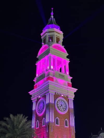The image shows the Clock Tower in Phú Quốc, Vietnam, at night. The tower is lit with purple and pink lights, which illuminate the building's architecture, including its arched windows, clock faces, and decorative trim. The clock faces show different times. A palm tree is visible at the bottom right corner of the image. Two beams of blue light extend from the top of the tower into the dark night sky.