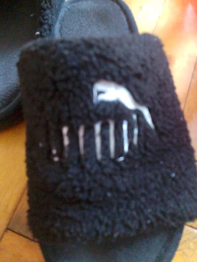 This is a close-up, slightly blurry, vertical photograph of a black, fuzzy Puma slipper. The slipper is the primary object, positioned centrally and taking up most of the frame. The material appears soft and plush, with a textured surface.
On the front of the slipper, there is a white Puma logo. The logo is partially obscured by the fuzzy texture and the blurriness of the image, but the characteristic leaping cat shape is discernible. Below the cat logo, there are several vertical white lines, which may be part of the slipper's design or perhaps some residue or stain. There are also a few small, scattered white specks on the slipper's surface.
The slipper is resting on a wooden floor, which is visible in the background, particularly on the right side of the image. The floorboards have a warm, orange-yellow hue, and the lighting suggests a bright environment, possibly an indoor setting with natural light coming from a window.
No people are visible in the photograph. The activity is static; the slipper is simply presented. The notable details are the slipper itself, its brand, and its material. There is no information about the time of day, weather, or specific emotions conveyed. The location context provided is Melitopol, Ukraine, but there are no visual cues within the image to confirm this specific location. No visible text other than the brand logo is present.