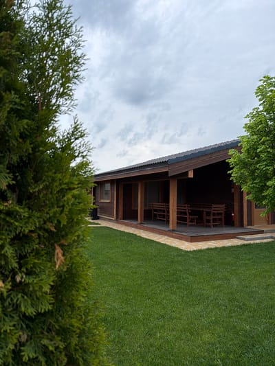 The image depicts a single-story wooden building in a green, grassy environment, likely taken in a rural or suburban area. The building has a covered porch area furnished with wooden chairs and what appears to be a fire extinguisher mounted on the wall. The sky is overcast. A coniferous shrub occupies the foreground. The image does not provide explicit evidence of its location in Kamyshin, Russia. Without additional visual markers or location tags, confirmation is impossible.