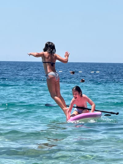 In this image, two children are engaged in watersports on a bright, sunny day. A young girl in a striped bikini is in the foreground, her body extended horizontally as if diving or falling into the water. Her arms are outstretched, and her legs are together. Behind her, slightly to the right, a boy wearing swim trunks is on a paddleboard. He is holding a paddle, which has a pink floatation device attached to it. The paddleboard is positioned in the turquoise water, which is clear enough to see the sandy bottom beneath. The scene appears to be set in a calm bay or lagoon, with the vast blue sky and the shimmering surface of the water creating a serene atmosphere. The water is clear and reflects the sunlight, creating a beautiful, rippling effect. In the background, several other people are visible in the water, some with their heads above the surface, suggesting a popular recreational spot. The overall mood is one of carefree summer fun and aquatic adventure. The location is identified as Mirište, Montenegro. There is no visible text in the image.