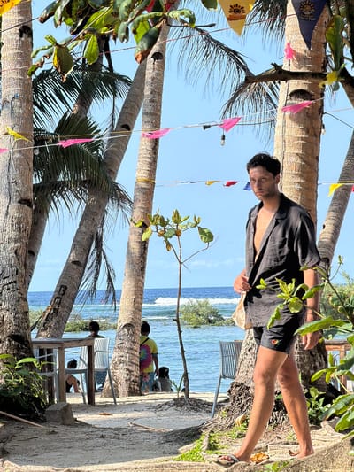 The image shows a beach scene in General Luna, Philippines. A man in a black shirt and shorts stands in the foreground. In the background, several people are sitting at tables near the water. Palm trees and colorful flags are present, creating a tropical atmosphere.