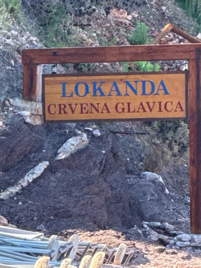A weathered wooden signpost stands in a rocky, outdoor environment. The sign itself is wooden and rectangular, with two lines of text. The top line reads "LOKANDA" in blue capital letters. The bottom line reads "CRVENA GLAVICA" in maroon capital letters. The signpost is supported by a dark brown wooden frame, with vertical posts and a horizontal beam connecting them. In the background, there is a rocky hillside with sparse vegetation. In the foreground, there are some metal structures and rocks. The overall impression is that of a rustic sign marking a location in a natural setting.