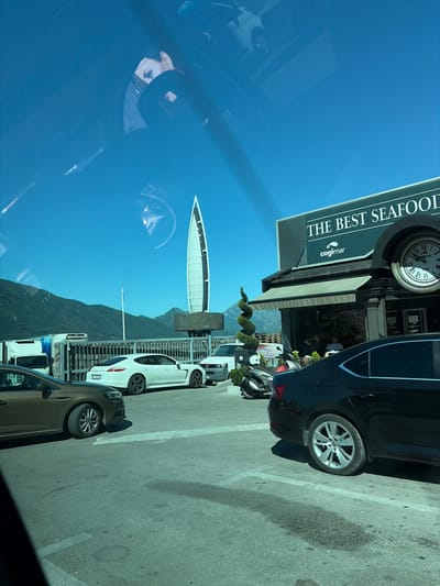 A reflection of a woman's face is visible in the upper portion of the image, superimposed over a bright blue sky and a distant mountainous landscape. Below the sky, a modern building with the prominent text "THE BEST SEAFOOD" and a logo featuring a fish is seen. A large clock with Roman numerals is mounted on the building. In front of the building, there are several parked vehicles, including a white Porsche Panamera, a dark-colored sedan, and a white truck. A spiral-shaped topiary is also visible in front of the building. The scene appears to be a street or parking lot in front of a seafood restaurant. The license plate of the white Porsche reads "KO AB524". The environment suggests a sunny day in a coastal town. The location is Dobrota, Montenegro.