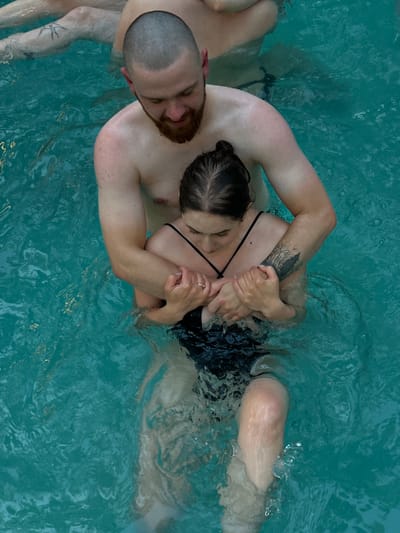 In Ivachiv Horishnii, Ukraine, two individuals are submerged in turquoise water. A man with a shaved head and beard, shirtless, embraces a woman from behind. She wears a black swimsuit and has her hair pulled back. Their hands are clasped around her chest. The water appears to be a pool or similar body of water.