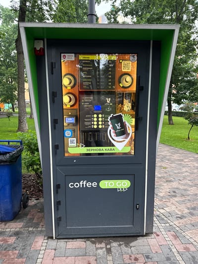 A dark gray "coffee TO GO" vending machine with a green and white angled canopy stands on a red and gray patterned brick pathway in Irpin, Ukraine. The machine features a prominent central display screen showing a menu in Ukrainian, offering drinks such as Espresso, Americano (with or without milk), Latte, Cappuccino, Mochaccino, Chocolate, and Tea with lemon, priced between 33 and 48 UAH. Payment options include a contactless NFC reader and multiple QR codes for digital transactions. Text on the screen indicates "BEAN COFFEE" (ЗЕРНОВА КАВА) and provides instructions (Place cup, Choose drink, Pay). Security warnings in Ukrainian ("ATTENTION! object under protection" with a padlock icon, and "TECHNICAL SUPPORT" with a CCTV icon) are visible, along with a small red and white alarm device mounted on the upper left side of the machine. A graphic depicting a hand holding a "12 coffee" branded cup acts as a point-of-interest marker on the screen. To the immediate left of the machine, a blue plastic wheeled dumpster with a black liner is positioned on a patch of dark mulch and green grass. The surrounding environment consists of green lawns, mature trees, and distant residential buildings under an overcast sky.
