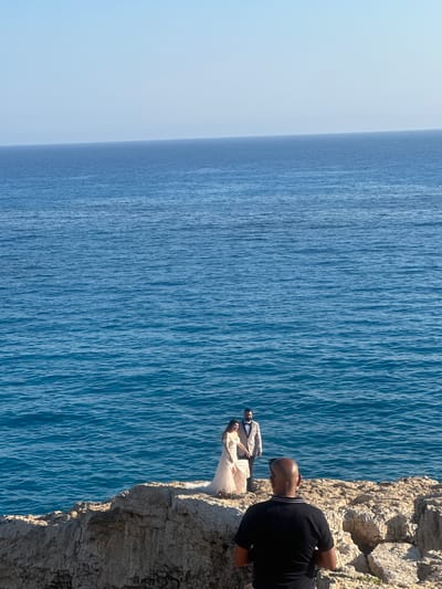 The image depicts a couple dressed in formal attire, a woman in a wedding gown and a man in a suit, posing for photographs on a rocky cliff overlooking the Mediterranean Sea. The sea is a deep blue, with gentle waves. The sky is clear and light blue. In the foreground, a man, possibly a photographer, is visible from behind, facing the couple. The location is identified as Ayia Napa, Cyprus, a popular tourist destination known for its beaches and natural beauty. The overall scene suggests a wedding photoshoot or a celebratory occasion in a scenic coastal setting.