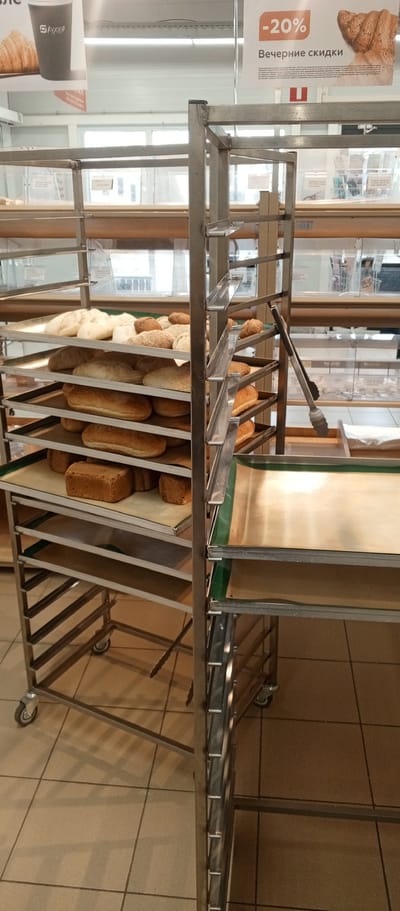 The image depicts a bakery display rack filled with various types of bread. The rack is made of stainless steel and has multiple shelves, each lined with baking mats. On the shelves, there are loaves of white bread, brown bread, and smaller bread rolls. Some bread is sliced, while others are whole.

In the background, there are signs. One sign, partially visible, has Cyrillic text and a "20%" discount offer with an image of bread. Another sign shows a coffee cup with the logo "М Кухня" (M Kuhnya), suggesting a cafe or bakery setting. The lighting suggests it is daytime. The floor is tiled with light-colored square tiles.

The scene appears to be inside a retail establishment, likely a bakery or a supermarket with a bakery section, in Zhirnovsk, Russia. The overall impression is a well-stocked display of fresh baked goods. There are no people visible in the frame. The focus is entirely on the bread and the display equipment.