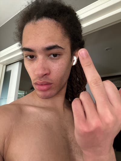 A rotated selfie captures a close-up of a person, likely male, with medium-dark skin, dark curly hair, and light facial stubble. The individual is shirtless, revealing a bare chest. In their left ear, a white earbud (resembling an Apple AirPod) is visible. The most prominent feature is the person's right hand, positioned prominently in the upper right quadrant of the frame, with the middle finger extended directly towards the camera, conveying a defiant or insulting gesture.

The person's head is tilted, with their eyes looking directly at the camera. Their facial expression is relatively neutral, but the hand gesture strongly dictates the confrontational tone of the image. The background consists of indoor architectural elements: a light-colored wall, white molding or a door frame, and a grey ceiling, suggesting an interior setting. The lighting appears to be artificial indoor light. There is no visible text in the image. The photograph captures a direct, provocative interaction with the viewer, taken in an indoor setting, potentially in Les Ulis, France.