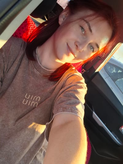 A woman with reddish-brown hair is taking a selfie from inside a vehicle. She is looking at the camera with a slight smile. Sunlight is streaming in from the side, illuminating her face and hair. She is wearing a tan, stone-washed t-shirt with a subtle embroidered logo on the chest, which appears to be stylized letters that resemble "UIM". The background shows the interior of a car, including a door panel with a silver handle and a window. The seat next to her is covered with a bright red, textured fabric. The lighting suggests it is daytime, likely late afternoon given the warm, angled sunlight. The overall mood is casual and relaxed. There is no information in the image to suggest a specific event or activity beyond a selfie being taken during a car ride. The location is provided as Kamyennoye, Russia.