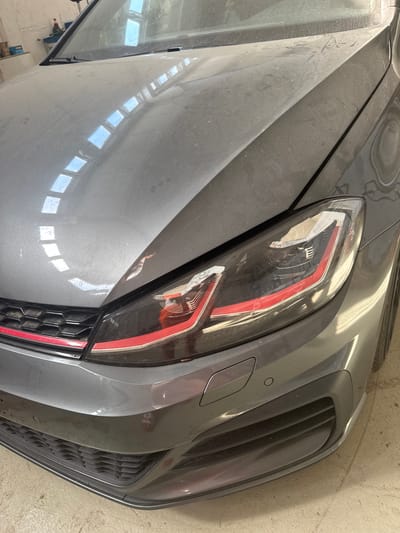 The image shows a close-up, high-angle view of the front right side of a dusty, dark gray car, possibly in a garage or workshop in Schkopau, Germany. The car's headlight, with its distinctive red and white accents, is a prominent feature. The headlight lens is slightly smudged, consistent with the overall dustiness of the vehicle. Next to the headlight, a portion of the car's grille is visible, featuring a honeycomb pattern and a thin red accent strip. Below the grille, a circular parking sensor and a rectangular cover for another sensor or feature are visible.

The car's body panels are visible, including the hood and fender, both covered in a layer of dust. The reflection of several rectangular windows or light fixtures can be seen on the car's hood, suggesting an indoor setting with overhead lighting. The background shows a concrete floor and a wall, further supporting the idea of an indoor location. No people are visible in the image. The lighting appears to be artificial, typical of an indoor environment. The overall impression is of a car that has been sitting idle, accumulating dust.