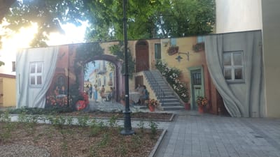 This is a mural painting on a building's exterior wall, depicting a lively outdoor scene in what appears to be a European town square. The mural is rich in detail, featuring architectural elements like arched doorways, staircases, and windows, all rendered with a sense of depth and perspective.
In the foreground, a black lamppost stands prominently. The mural itself portrays several groups of people engaged in various activities. On the left, a couple is seated at a table, seemingly enjoying each other's company. To the right of them, under an archway, other individuals are seen standing and conversing, creating a social atmosphere. Further to the right, another couple is seated at a table, also interacting. The scene is further enlivened by painted potted plants and flowers, as well as a red scooter. A small dog is subtly depicted near a doorway on the far right, adding a touch of domesticity.
The mural creates an illusion of a real-life setting, with elements like draped curtains painted on the far right and left sides of the wall, making the artwork blend with the physical space. The background suggests a bright day, with sunlight filtering through trees overhead, casting dappled light. The overall mood conveyed is one of pleasant social interaction and idyllic town life. There is some faint text visible, likely an artist's signature or inscription, on the lower right corner of the mural. The location context provided is Łódź, Poland.