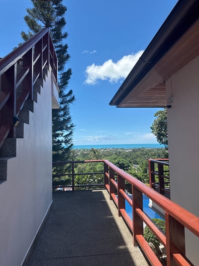 The image captures a view from a balcony or walkway in Ko Samui, Thailand, overlooking the ocean and lush green landscape. On the left, a white wall frames a granite staircase with a dark wooden railing. To the right, a similar dark wooden railing with red accents runs along the balcony, separating it from a blue swimming pool below. A white building with a wooden-paneled overhang and a security camera is visible on the right. A tall pine tree with dark green needles stands prominently between the staircase and the distant horizon. The sky is a vibrant blue with scattered white clouds. The overall scene suggests a tropical resort or residential setting with a scenic view.